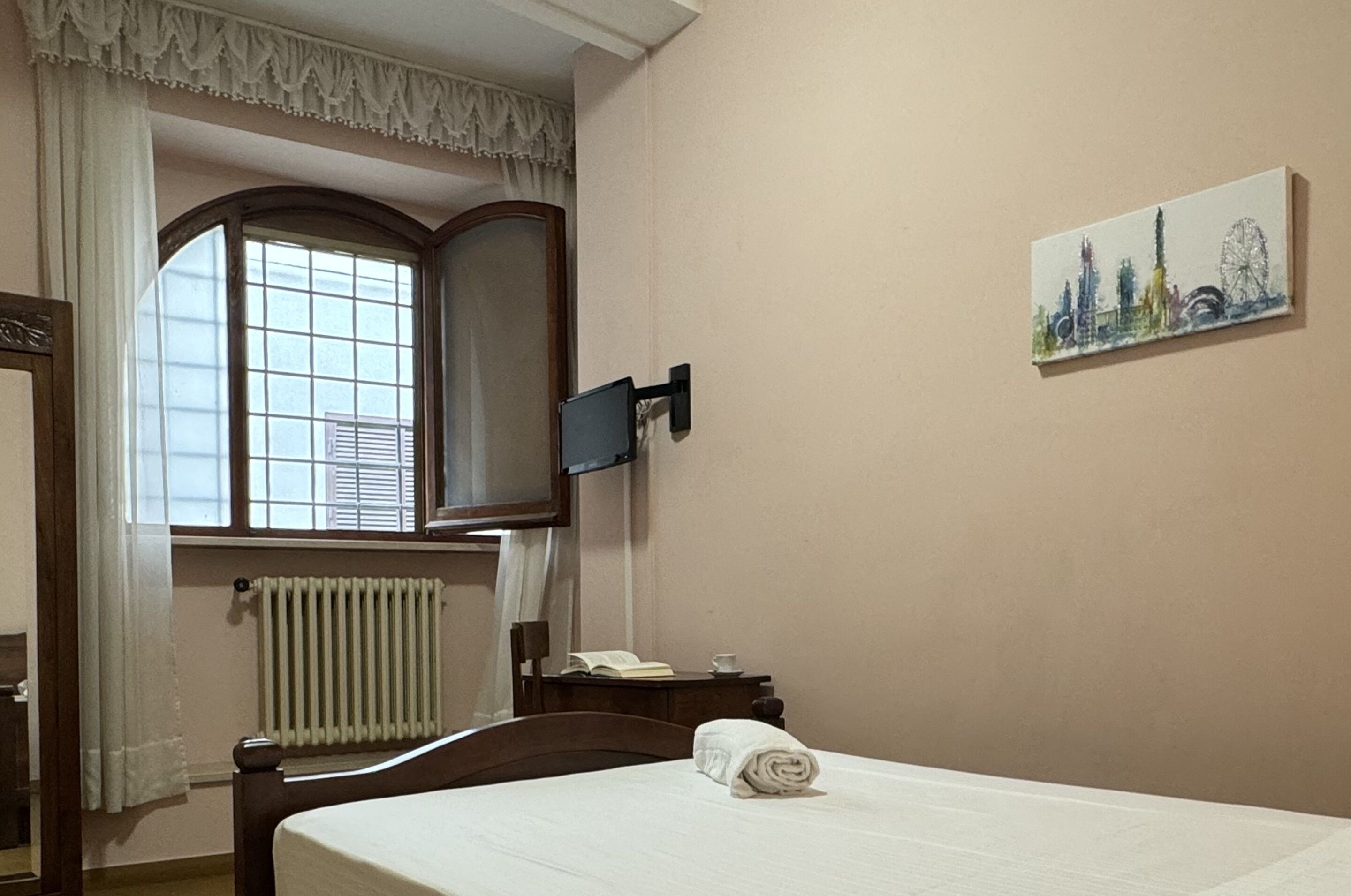 A hotel room with a bed, a window with curtains, a radiator, a wall-mounted TV, a small table with a book and a cup, and a painting on the wall.