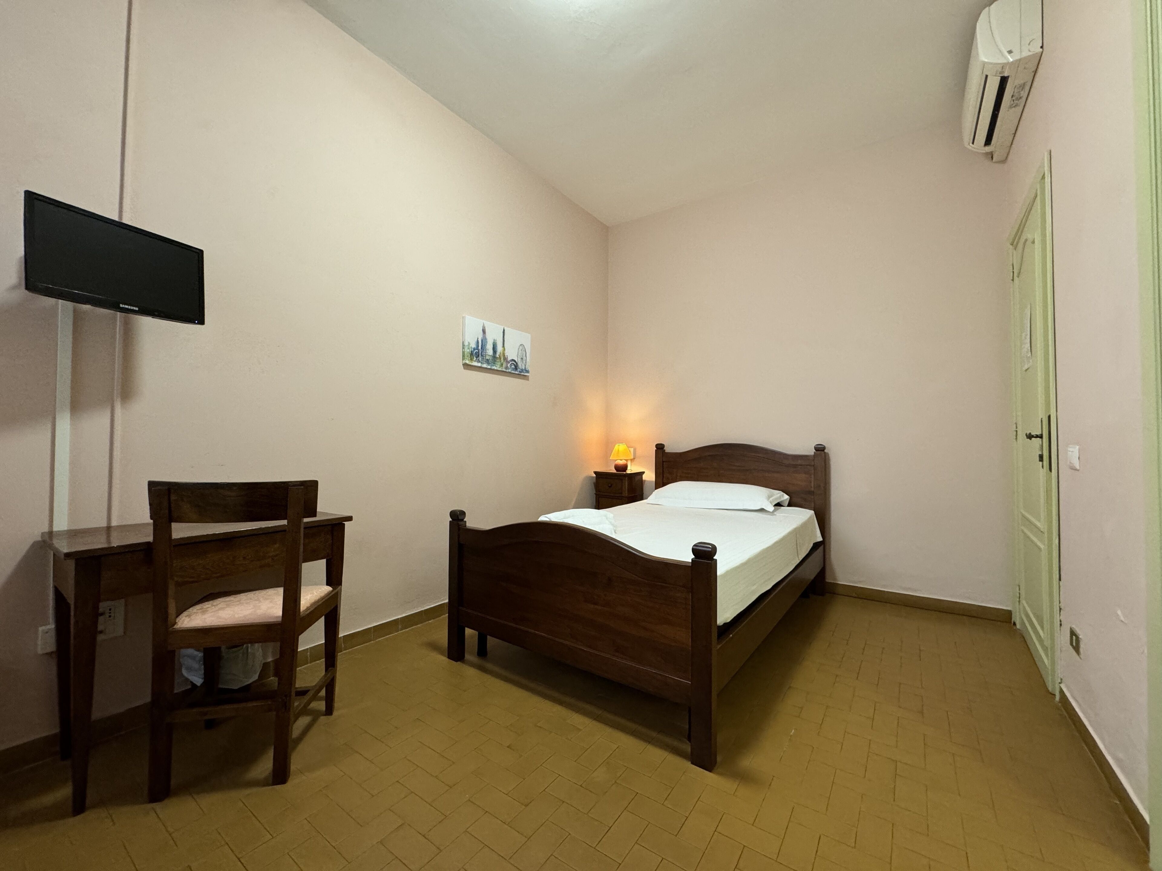 Um quarto com uma cama, escrivaninha, cadeira, televisão na parede, abajur e uma porta.