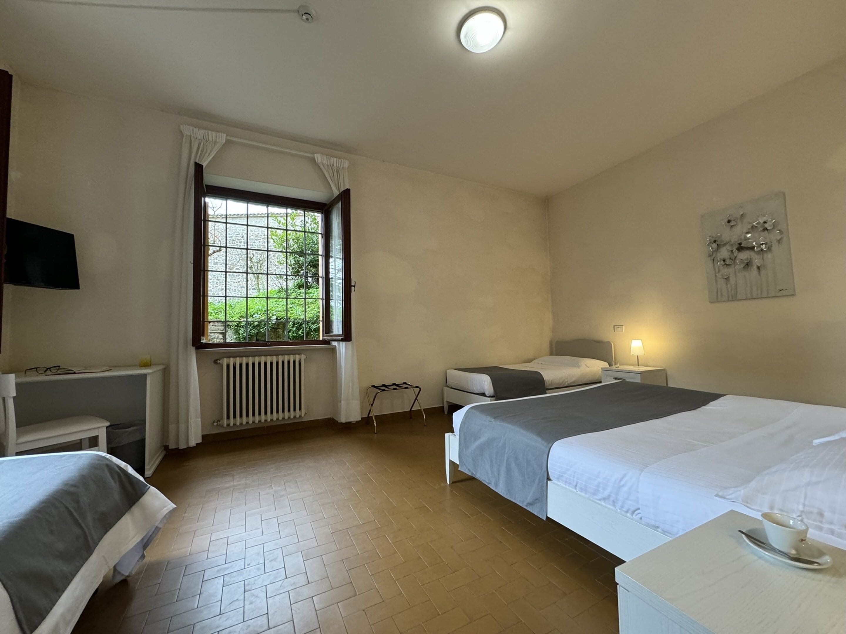 Une chambre d’hôtel avec deux lits, un radiateur, une fenêtre avec des rideaux, une table de chevet, une lampe et une œuvre d’art accrochée au mur.