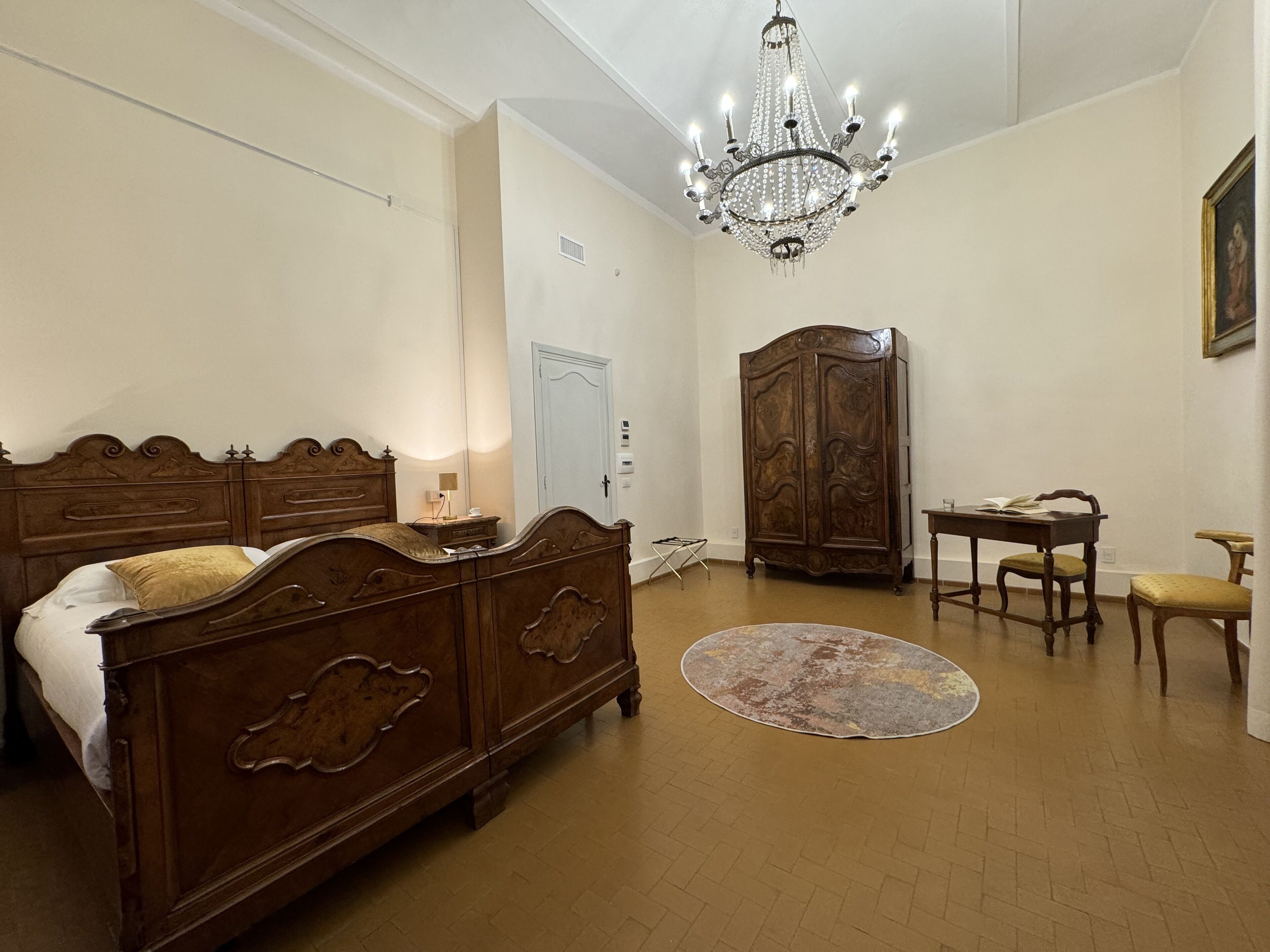 A bedroom with a wooden bed, a chandelier, a wardrobe, a desk with a chair, and a rug.
