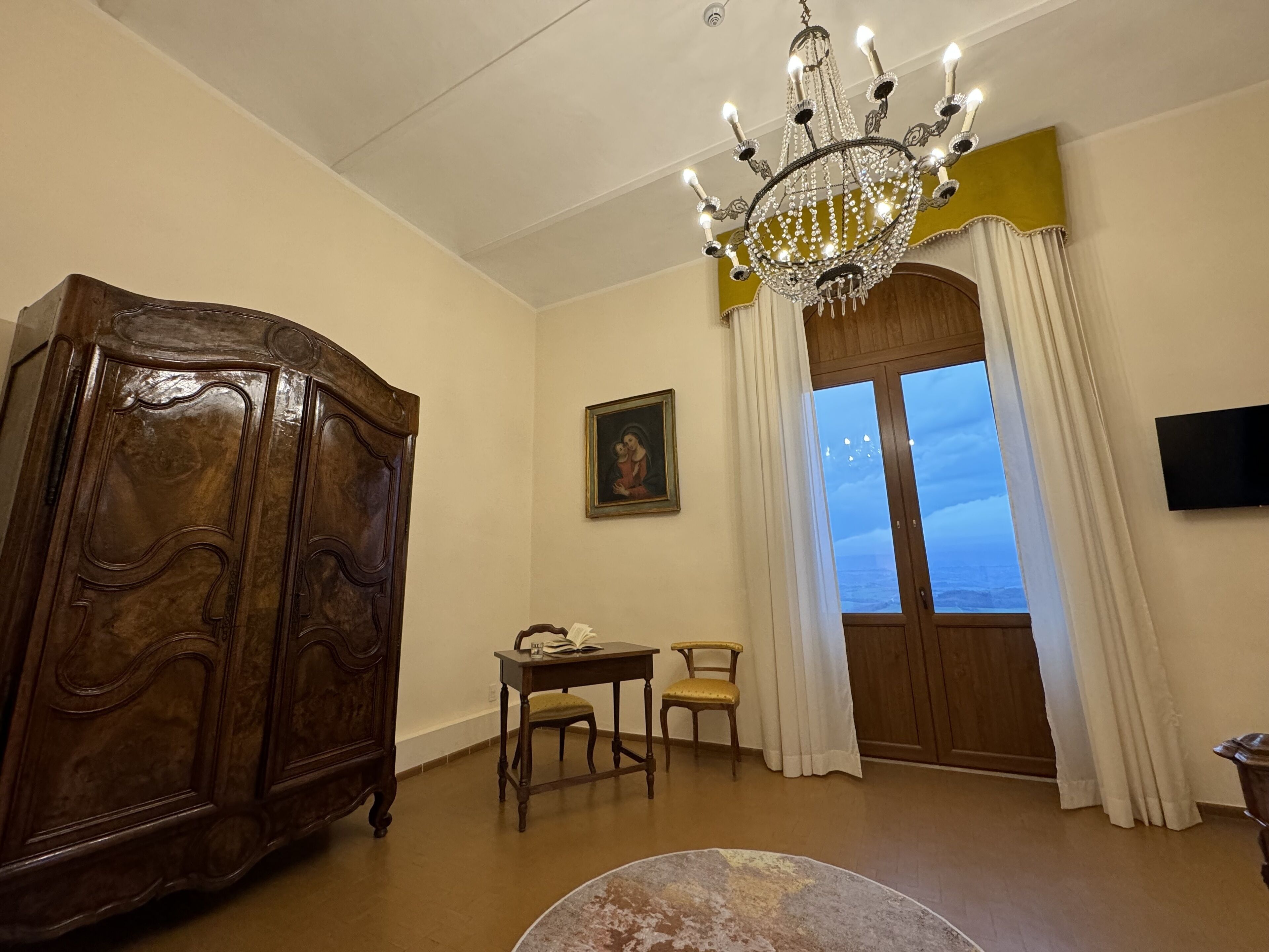 A room with a wooden cabinet, a table with a chair, a chandelier, a painting, and a window with curtains.