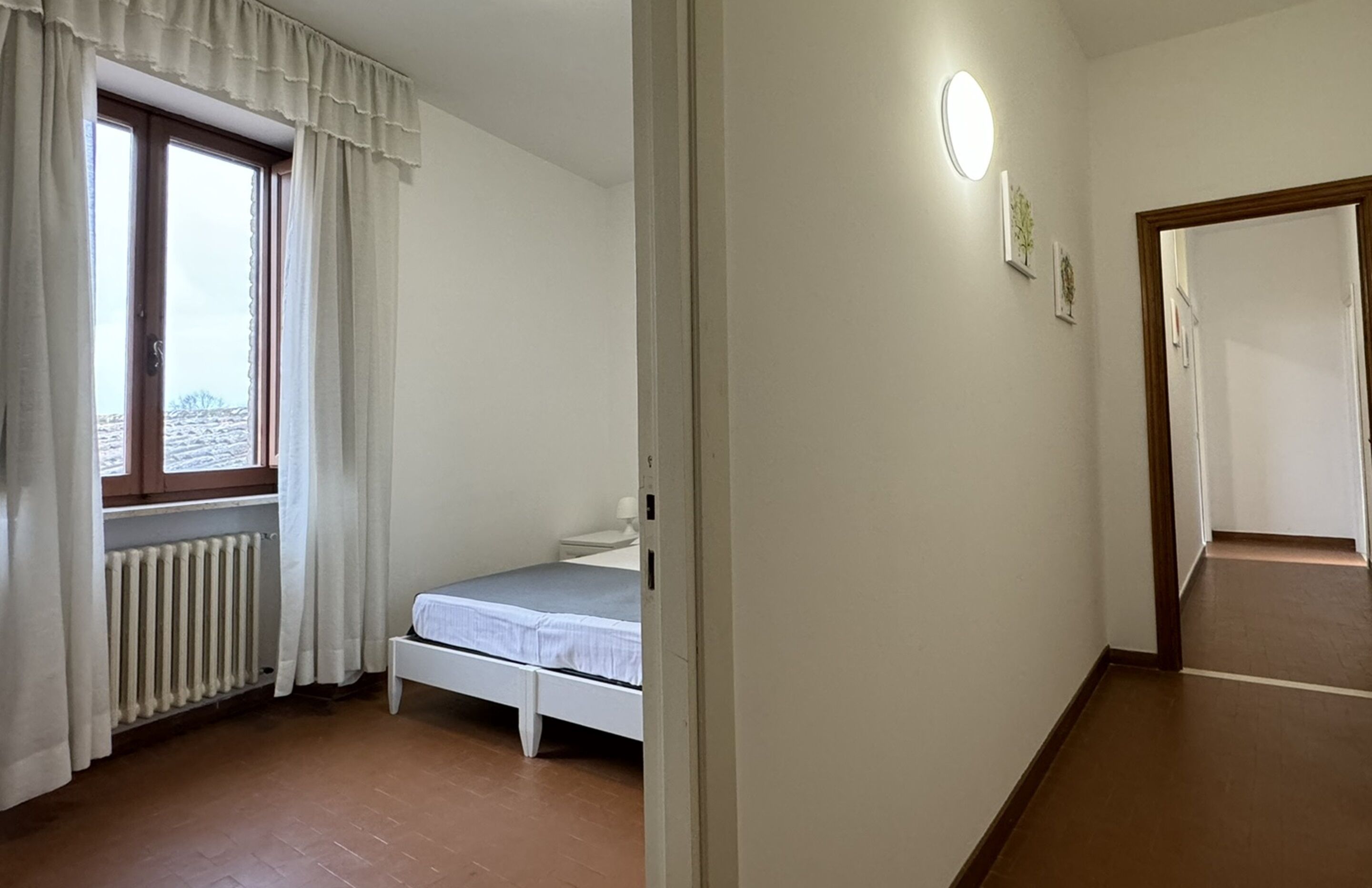 A small, single-bed room with a window, a radiator, and a wall-mounted light fixture.