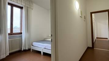 A small, single-bed room with a window, a radiator, and a wall-mounted light fixture.