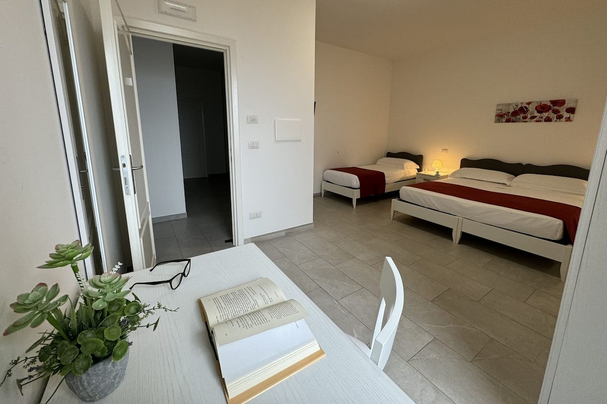 A hotel room with two beds, a desk with a book and glasses, a potted plant, and a door leading to another room.