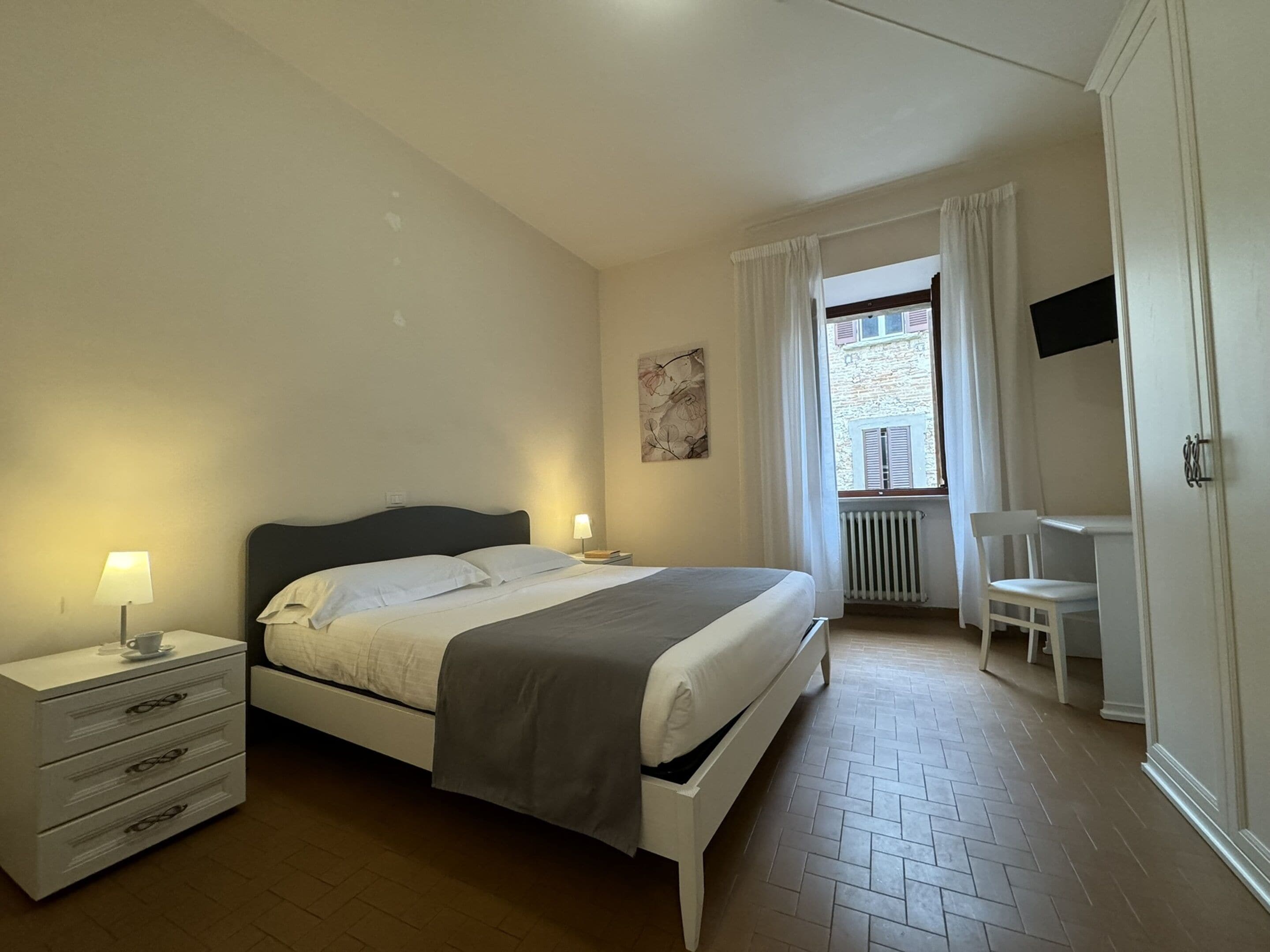 Ein Schlafzimmer mit Bett, Nachttischen, einem Sessel, einem Fernseher und einem Fenster mit Vorhängen.