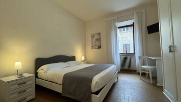 Ein Schlafzimmer mit Bett, Nachttischen, einem Sessel, einem Fernseher und einem Fenster mit Vorhängen.