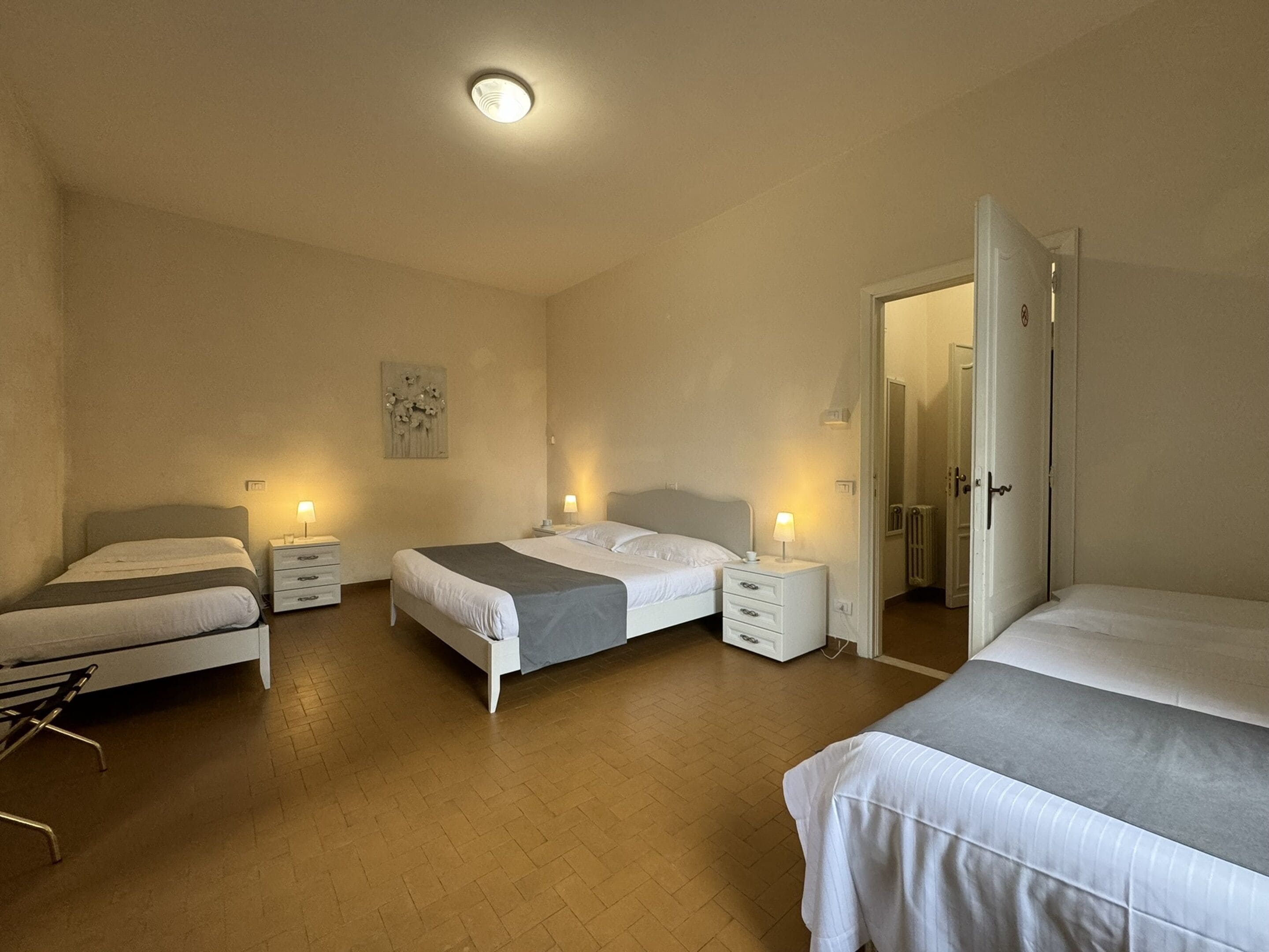 A hotel room with two beds, a chair, a bedside table with a lamp, a wall art piece, and a door leading to another room.