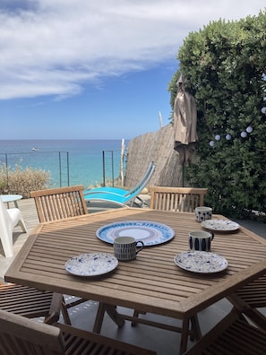 Um deck de madeira com uma mesa posta para refeições, com vista para uma piscina e o mar.