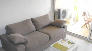 Un salón moderno con un sofá gris, una mesa de centro blanca y un amplio ventanal donde se ve un equipo de aire acondicionado.
