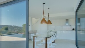 Un espace repas moderne avec une table en verre et des chaises blanches, une cuisine en arrière-plan et une vue sur l’extérieur.