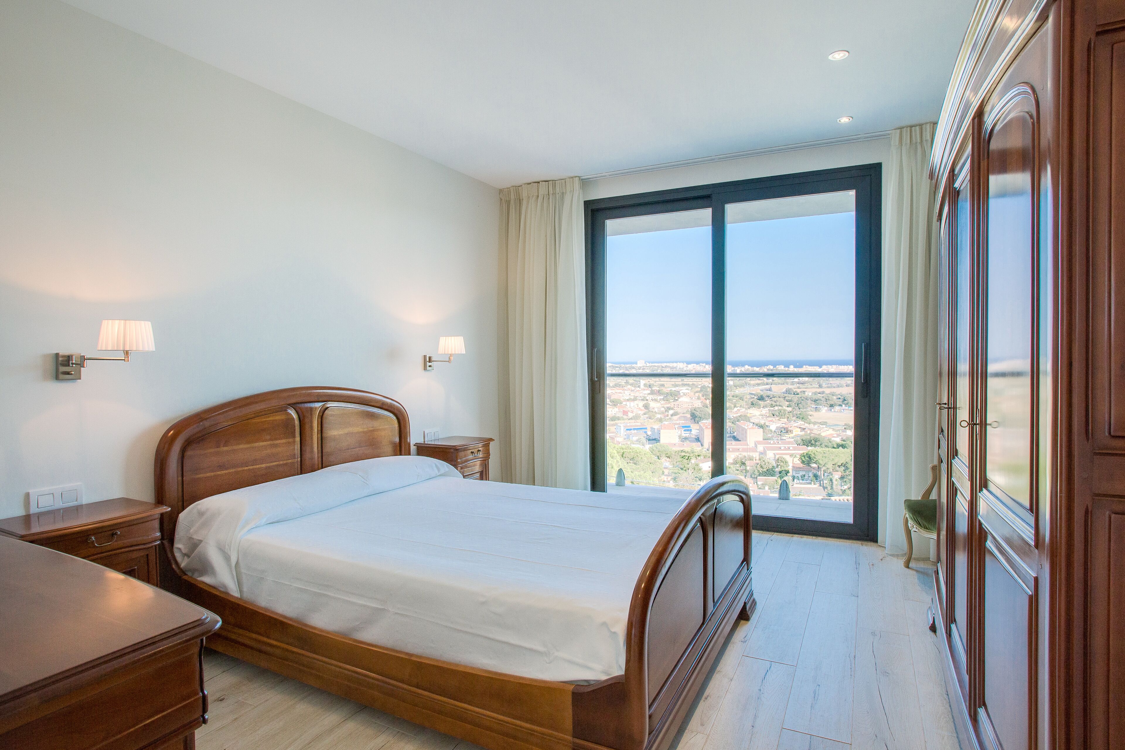 A bedroom with a large bed, a nightstand, a dresser, and a view of a cityscape through a large window.