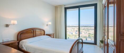 A bedroom with a large bed, a nightstand, a dresser, and a view of a cityscape through a large window.