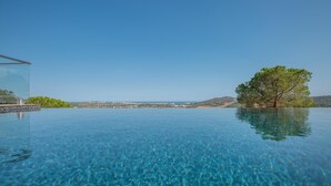 Une magnifique piscine à débordement d’un bleu profond, offrant une vue sur une côte lointaine et un arbre.