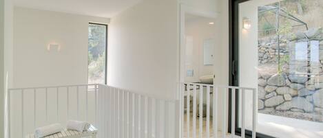 A modern, minimalist interior with a white bench, a glass door leading to an outdoor area, and a stone wall visible through the glass.