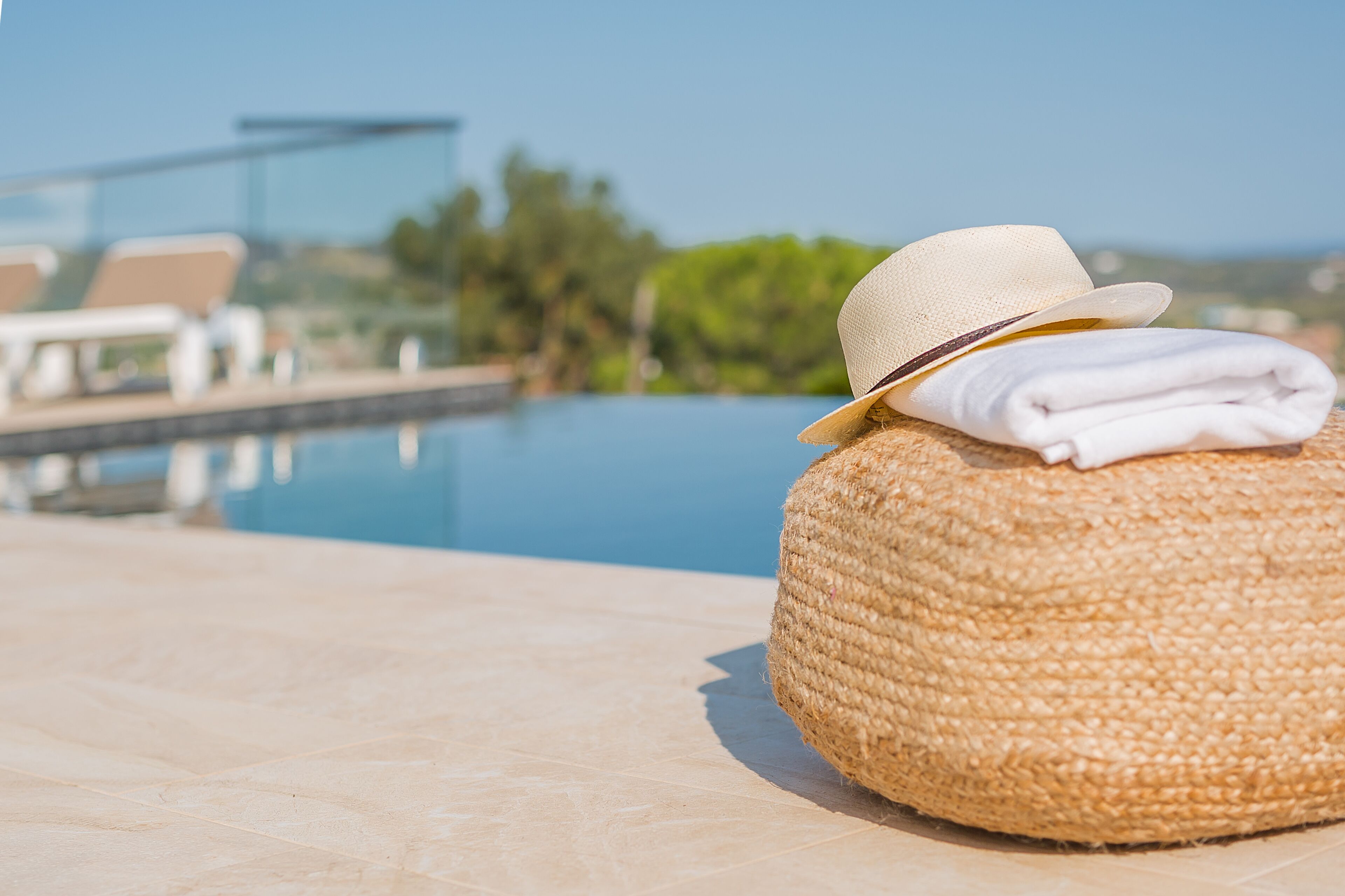 Un pouf rond en osier, sur lequel est posé un peignoir blanc plié et un chapeau beige, est placé au bord d’une piscine.