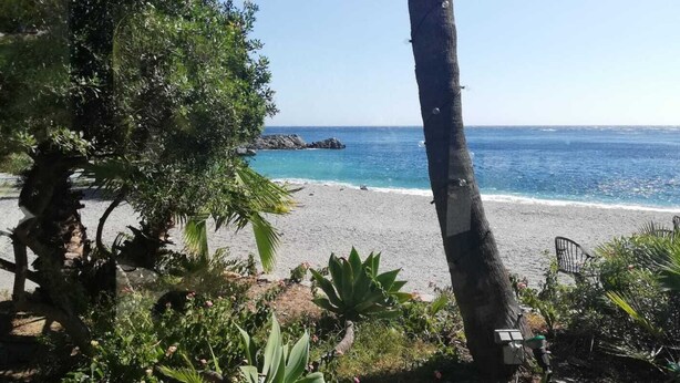 A beach with clear blue water, a rocky outcrop, and dense greenery.