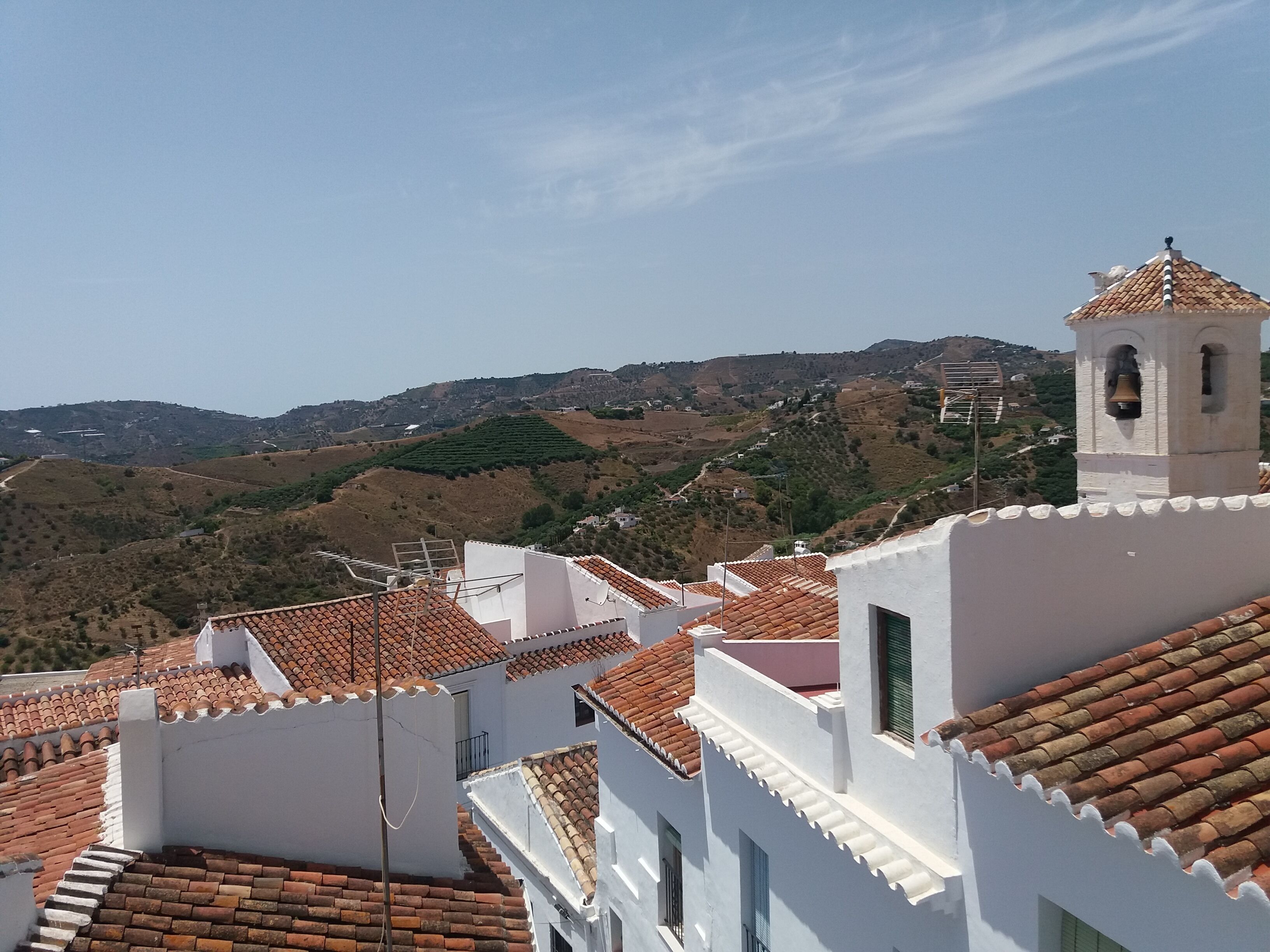 A view of terracotta-tiled rooftops and white buildings with a bell tower, set against a hilly landscape.