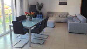 A modern living room with a glass dining table, black chairs, a grey sofa, and a grey armchair. There is a potted plant on the table and a painting on the wall. - Luxury  Repouso do Milhafre 181/AL (Horta)