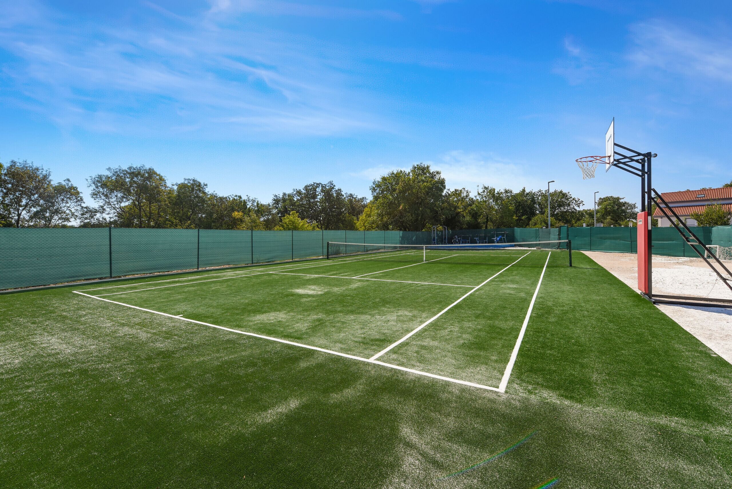 Cancha de tenis exterior con aro de baloncesto, rodeada por una valla verde y árboles al fondo.