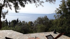 Um pátio de pedra com duas cadeiras de madeira e uma pequena mesa, com vista para um corpo de água e montanhas ao longe.