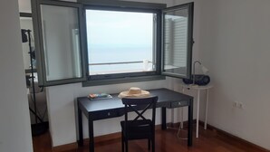Quarto pequeno com escrivaninha, cadeira e janela com vista para o mar.