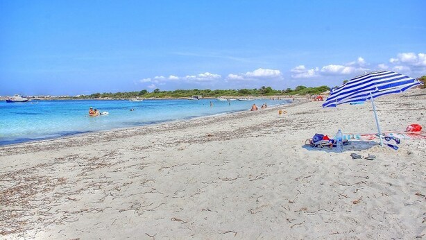 A beach with clear blue water, people swimming, and a striped umbrella on the sand.