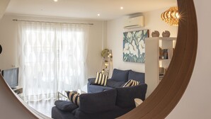 A modern living room with a dark blue sofa set, a large window with sheer curtains, a wall-mounted air conditioner, and a decorative light fixture.