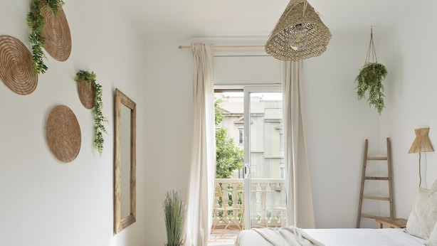 Une chambre à coucher avec un lit, un bureau en bois, une fenêtre avec des rideaux et un balcon agrémenté de plantes.