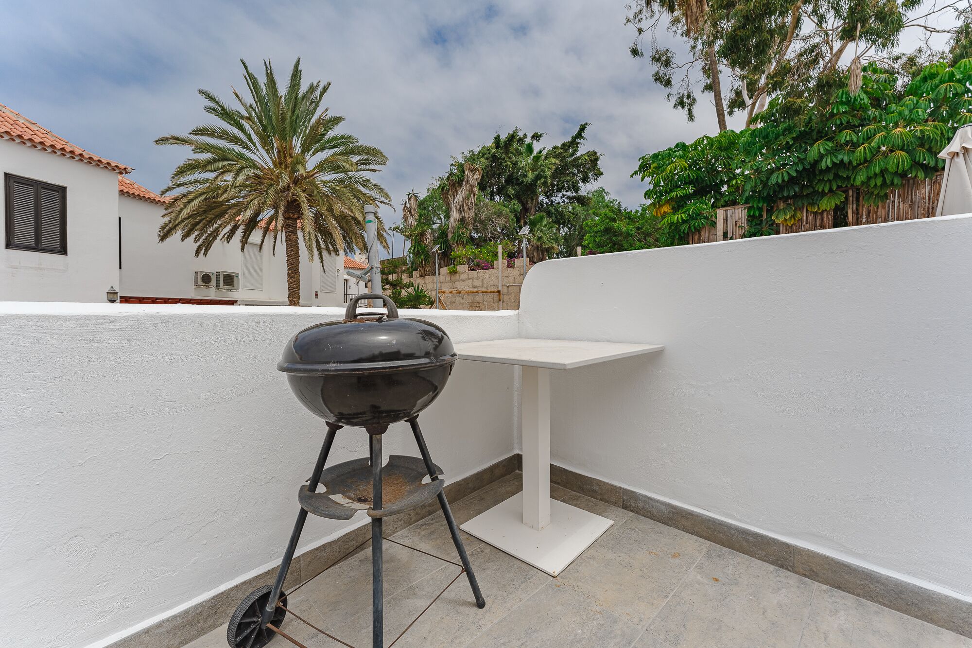 A portable barbecue grill on a balcony with a white wall, a palm tree, and a view of a building with a tiled roof.