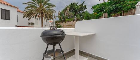 A portable barbecue grill on a balcony with a white wall, a palm tree, and a view of a building with a tiled roof.