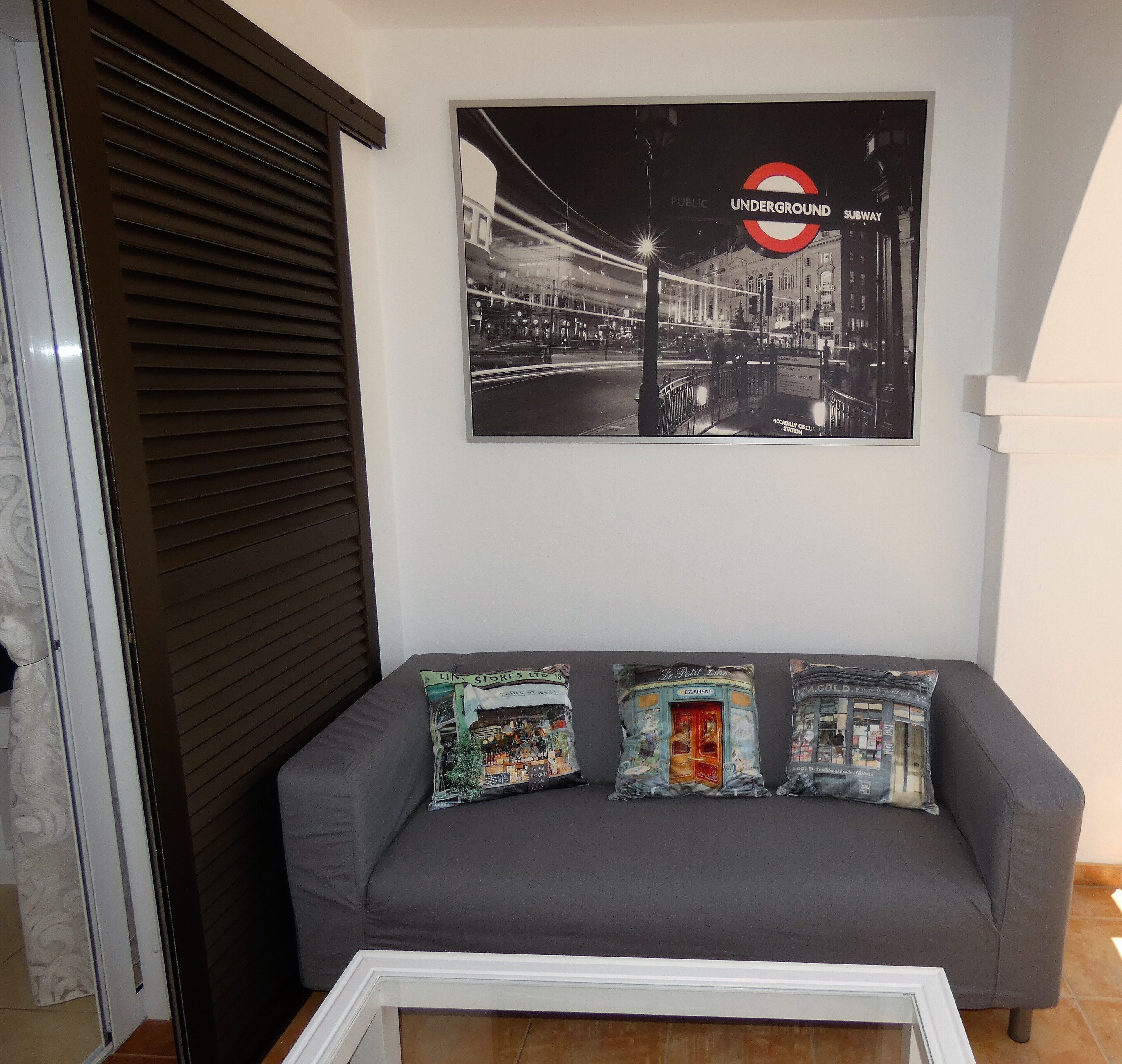 Un sofá gris con tres cojines decorativos, un cuadro enmarcado del metro de Londres y una ventana cerrada con persianas.