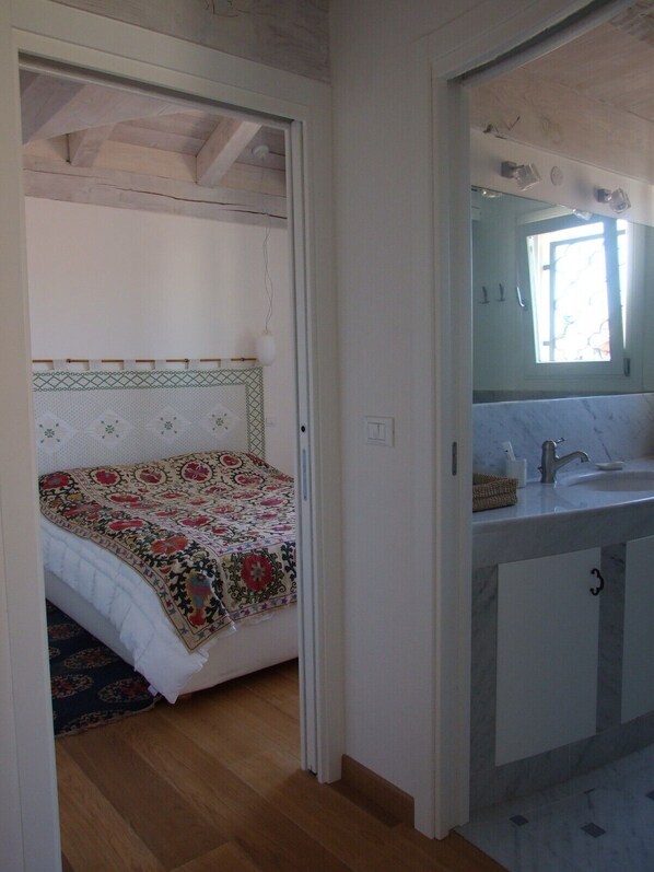 A bedroom with a bed, a bedside table, and a window. There is a bathroom with a sink and a mirror. - Stylish apartment 5 min walk from Rialto 360 degree panoramic view (venice)