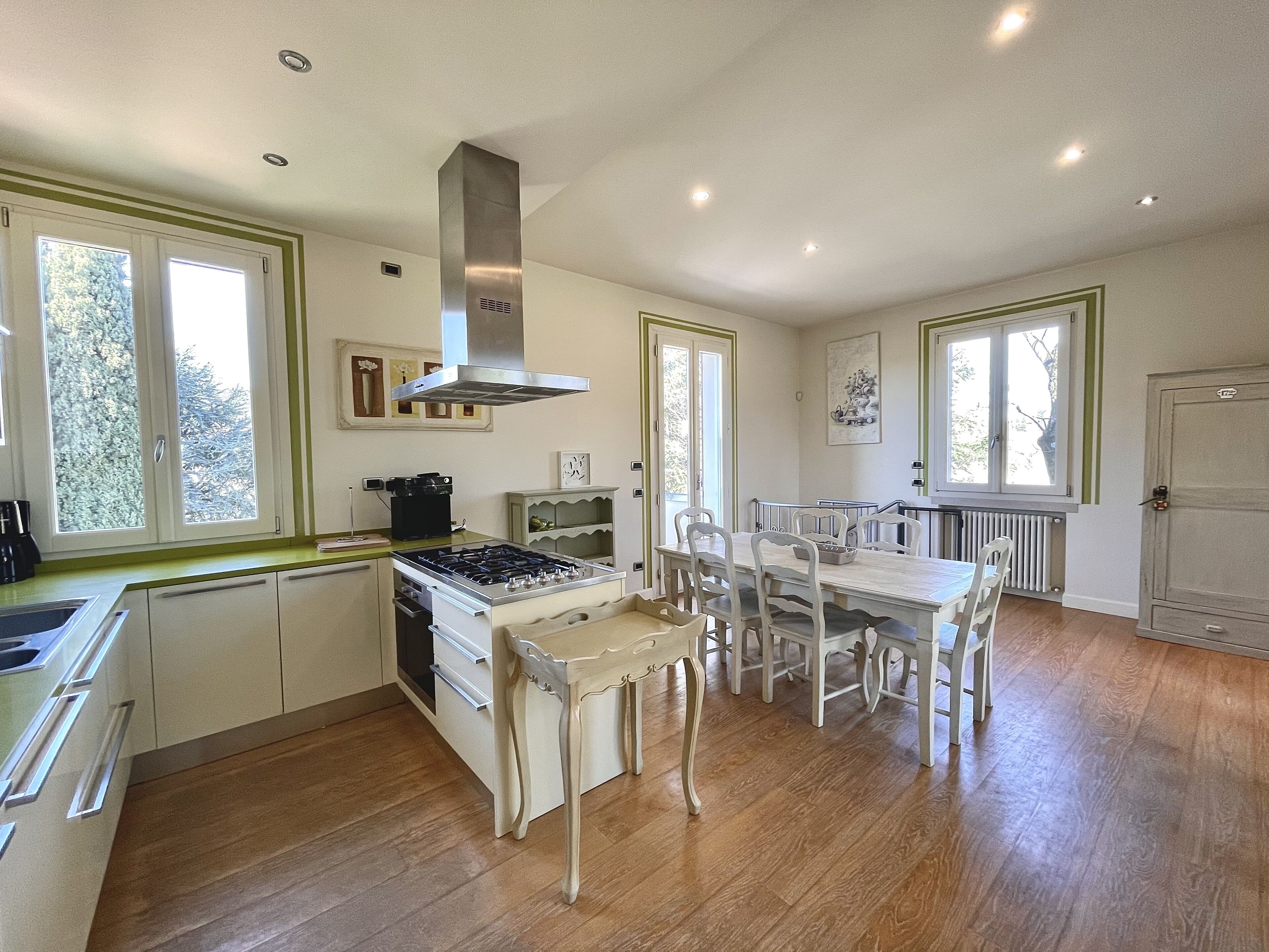 Una cocina moderna con armarios blancos, una estufa a gas y un extractor de acero inoxidable. Hay una mesa de comedor con sillas blancas y un ventanal con vistas a árboles.