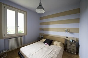 Une chambre à coucher avec un lit, une table de chevet, une lampe, une fenêtre et un radiateur.
