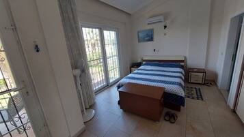A bedroom with a bed, a bedside table, a fan, and a window with a view of the outside.