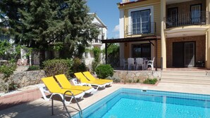 Ein Schwimmbecken mit gelben Liegestühlen, eine überdachte Terrasse mit Gartenmöbeln und eine Steinmauer.