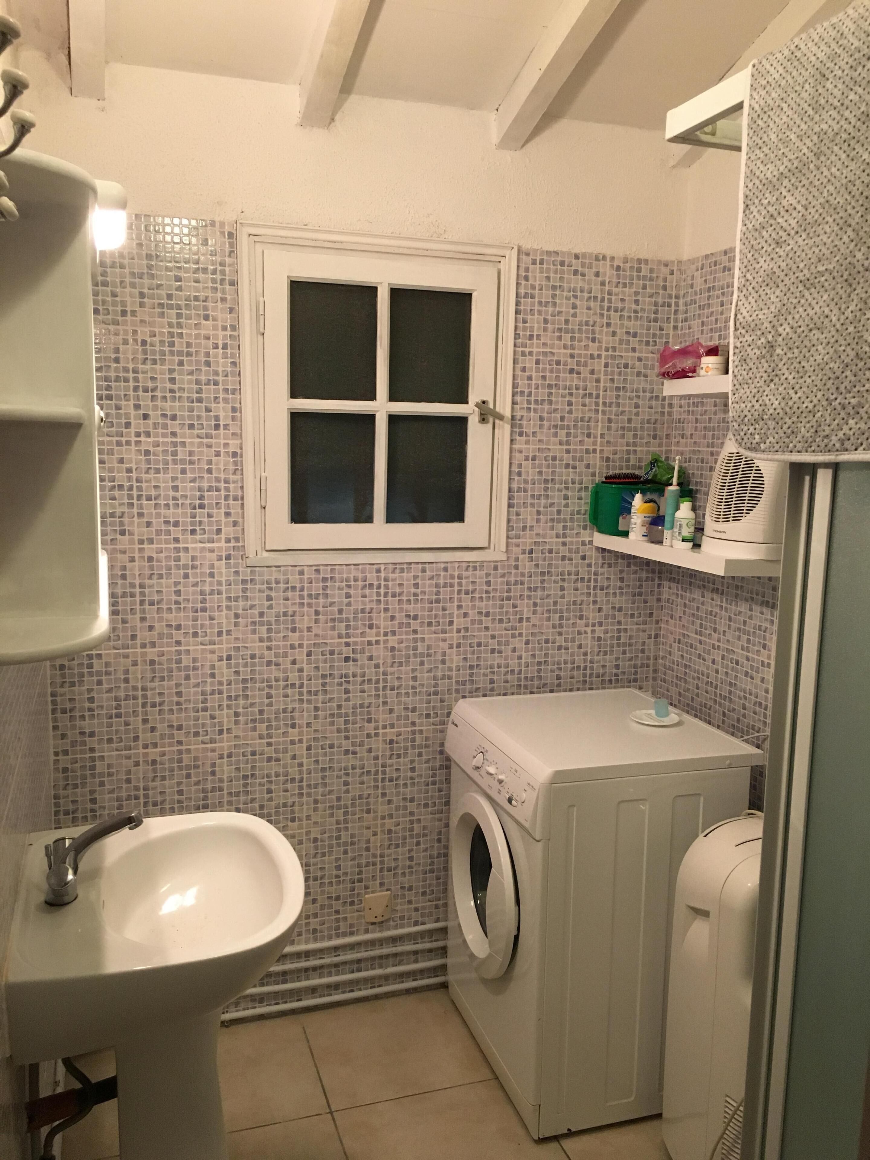A compact bathroom with a sink, a window, a washing machine, and a shelf with toiletries.