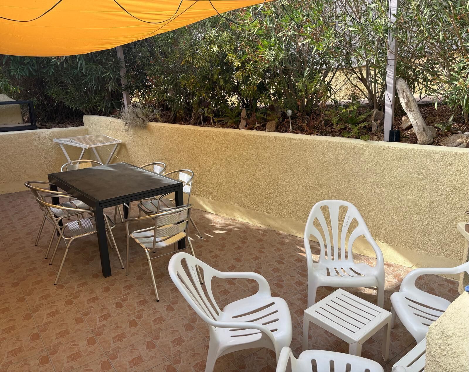 An outdoor patio area with a table and chairs, surrounded by a low wall and vegetation.