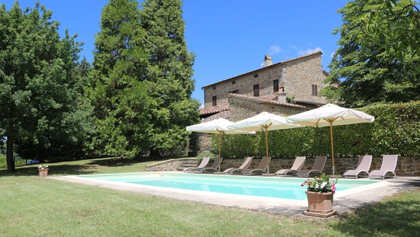 A swimming pool with lounge chairs and umbrellas, surrounded by greenery and a stone building in the background.