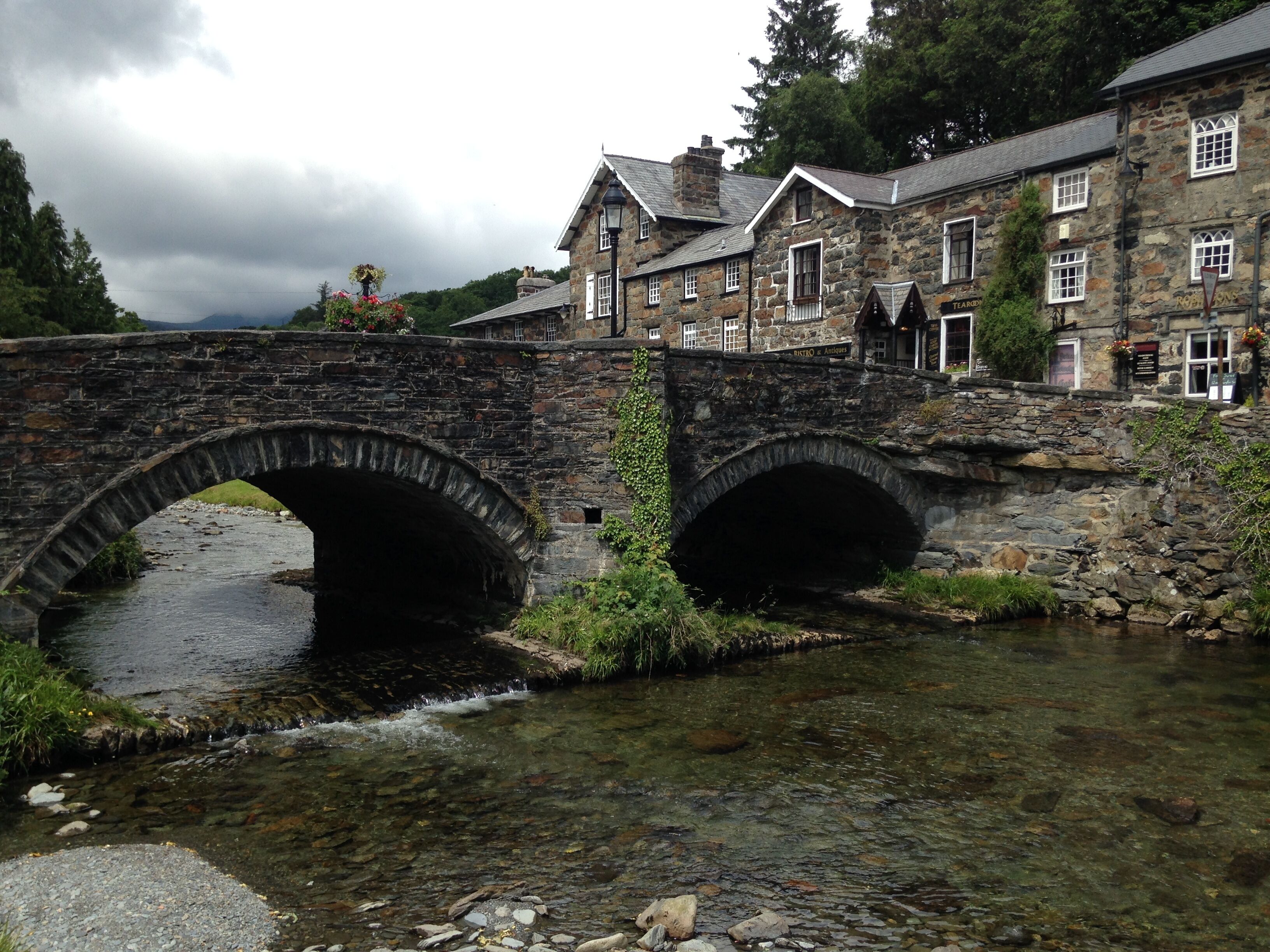A stone bridge over a clear stream with a building featuring multiple windows and a signboard in the background.