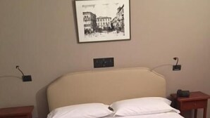 A neatly made bed with a quilted white duvet, a framed picture on the wall, and a small bedside table with a lamp. - Hotel il Cigno (Perugia)