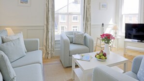 A modern living room with a grey sofa set, a white coffee table with a bowl of fruit and a magazine, a TV on a stand, and a view of buildings through large windows. - The Lighthouse - luxury hotel style, central apartment with fantastic sea views (Southwold)