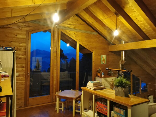 A cozy wooden cabin with a dining area, a table with books, a potted plant, and a view of the mountains through the glass door.