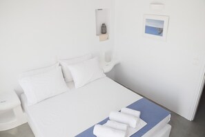 Ein ordentlich bezogenes Bett mit weißen Textilien und blauem Akzent, ein Nachttisch mit einer Lampe sowie eine kleine Ablage mit einer Vase und einem Bild.