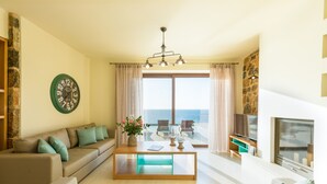 En moderne stue med sofa, sofabord og fjernsyn. Der er et stort vindue med gardiner, en stenvæg og udsigt til havet.