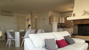 A modern living room with a white leather sofa, a dining area with white chairs, and a kitchen with wooden cabinets.
