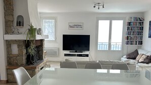 A modern living room with a glass dining table, white chairs, a stone fireplace, a flat-screen TV, and a bookshelf.