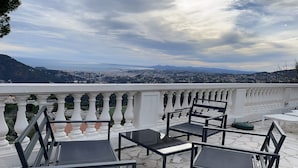 A terrace with outdoor furniture, a white balustrade, and a scenic view of a city and mountains in the distance.