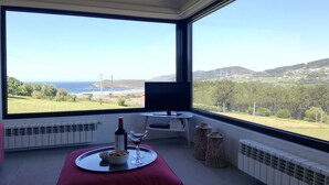 Une pièce dotée d’une grande fenêtre donnant sur un paysage côtier, une petite table avec une bouteille et un verre, et un pouf rouge.