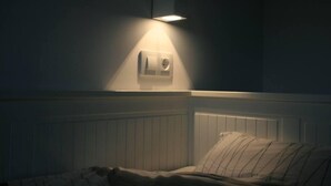 Une pièce faiblement éclairée, avec un lit, un luminaire fixé au mur et un thermostat.